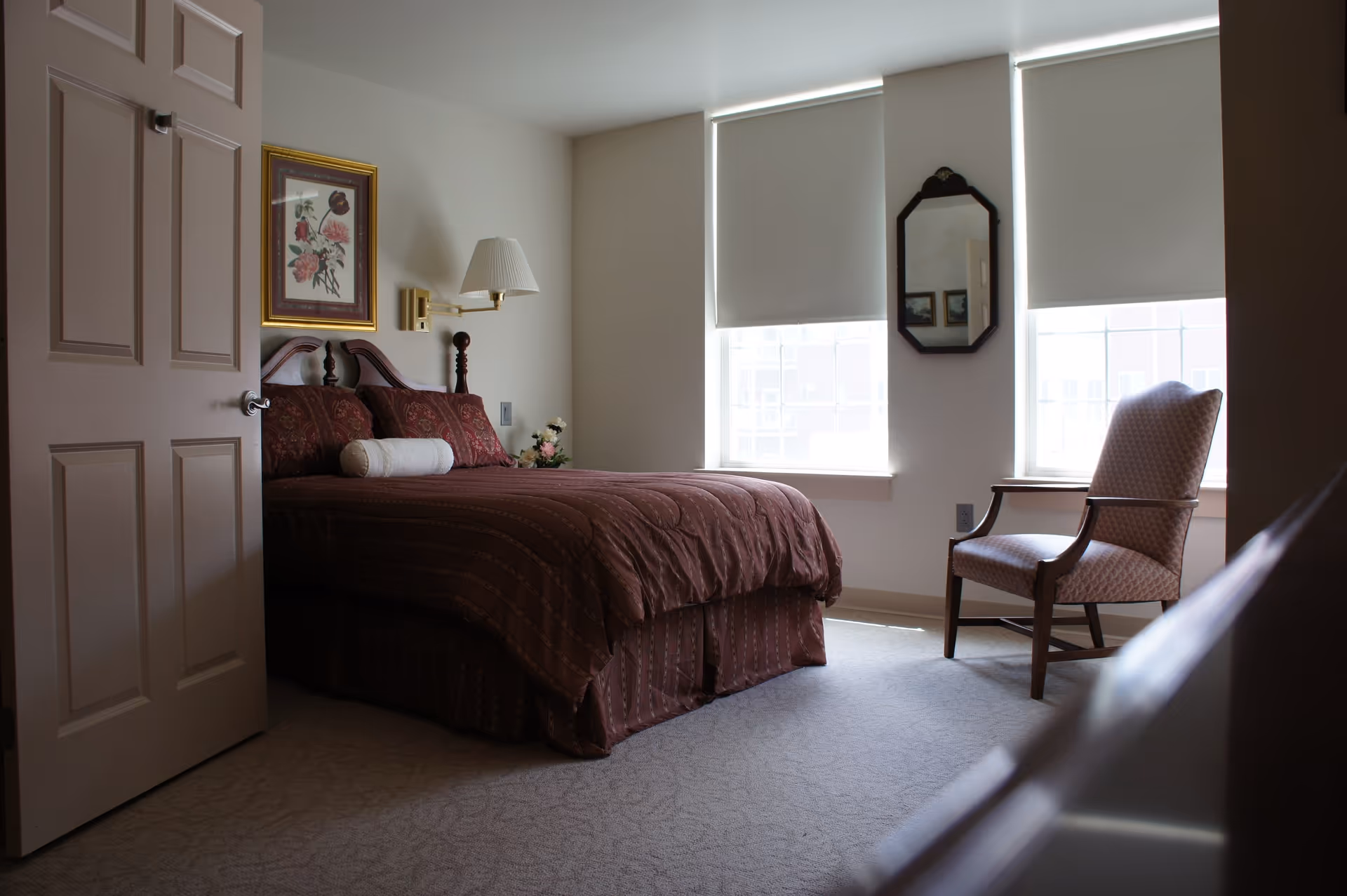 A bedroom with a bed covered in a dark red patterned comforter and pillows, a white bolster pillow, a floral painting on the wall above the headboard, a wall-mounted lamp, two windows with white roller blinds, a wooden framed mirror on the wall, and a cushioned armchair near the windows.