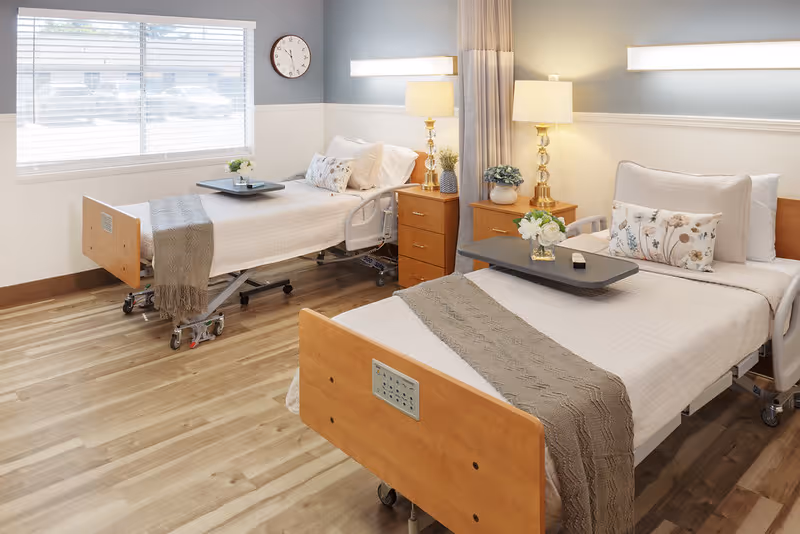 A bright and clean room with two hospital-style beds, each with white bedding, decorative pillows, and a beige throw blanket. Each bed has a small overbed table with a flower arrangement. Between the beds are two wooden nightstands with lamps and small plants. A large window with blinds lets in natural light, and a wall clock is visible above one bed. The floor is light wood, and the walls are painted in soft neutral tones.