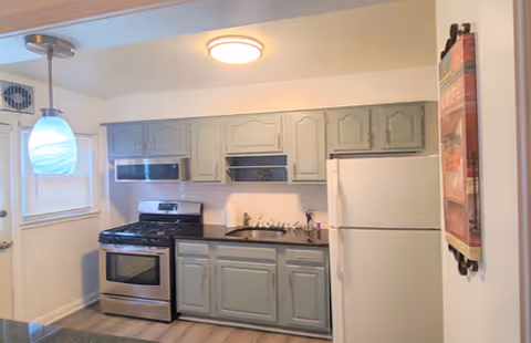 A kitchen area with light gray cabinets, a stainless steel gas stove with oven, a white refrigerator, and a black countertop. There is a small window with blinds on the left side and a hanging light fixture with a blue and white glass shade. A decorative towel hangs on the right wall.