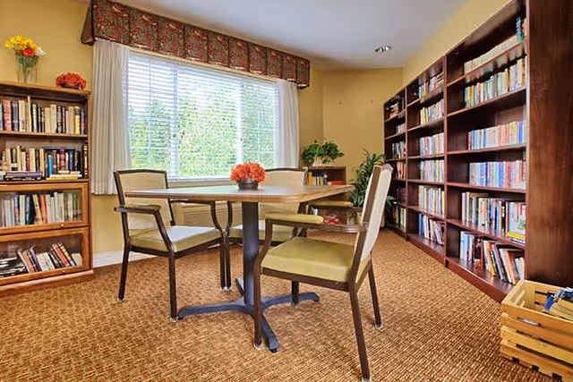 A cozy reading or sitting area with a round wooden table surrounded by four chairs with green cushions. The room has large windows with white blinds and a decorative valance, letting in natural light. There are tall wooden bookshelves filled with books on two walls, a small wooden crate with magazines or books on the floor, and a few plants and flowers adding a touch of color.