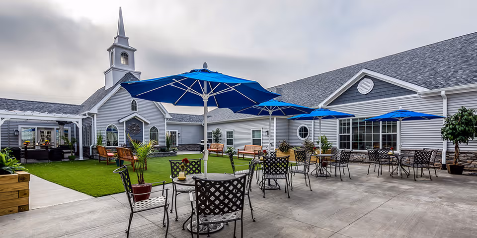 Outdoor courtyard with metal patio tables and blue umbrellas on a concrete patio beside a green lawn and a gray building with a small steeple.