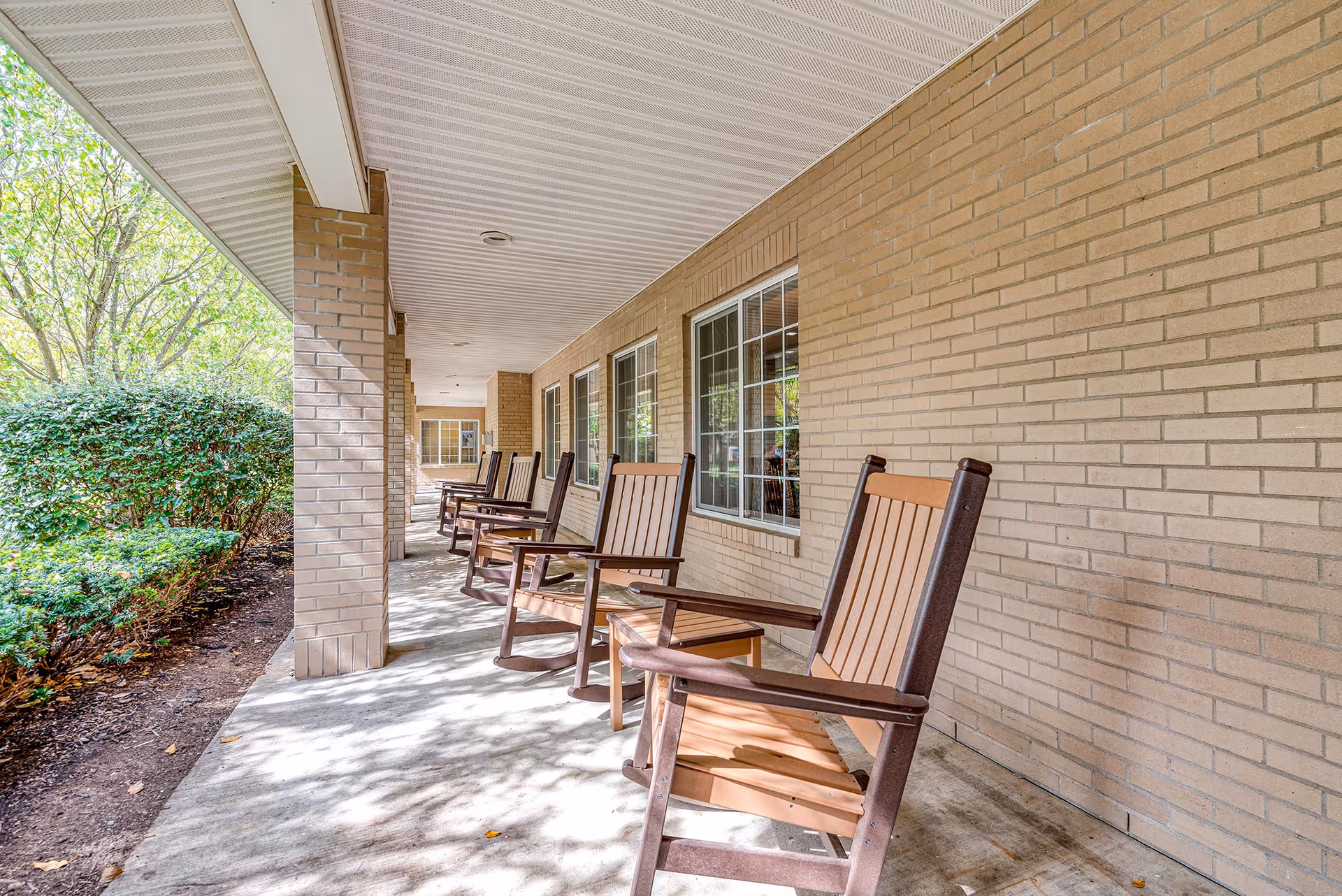 A covered brick porch with several wooden rocking chairs lined up facing a leafy garden.