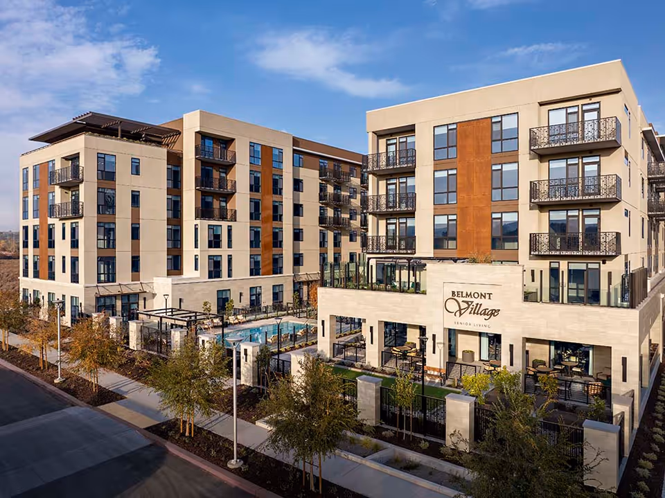 Exterior view of Belmont Village Senior Living San Ramon, showing a modern multi-story building with balconies, large windows, outdoor seating areas, and a swimming pool surrounded by trees and landscaping under a partly cloudy sky.