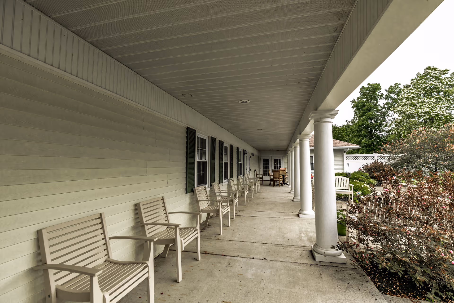 Covered porch with a row of white chairs and columns along the exterior of a senior living facility overlooking landscaped shrubs.