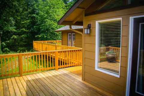 Wooden deck and railing extend from a tan building beside lush green trees.