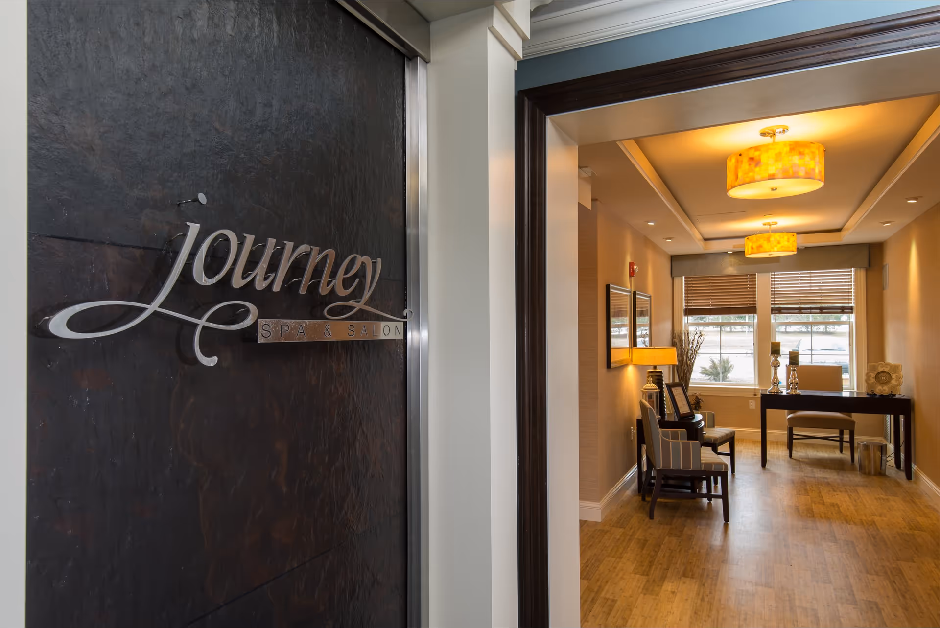 Entrance to Journey Spa & Salon with a dark textured door featuring the spa's name in metallic letters. Adjacent to the door is a warmly lit interior space with wooden flooring, two chairs, a table with decorative items, a lamp, and windows with blinds.
