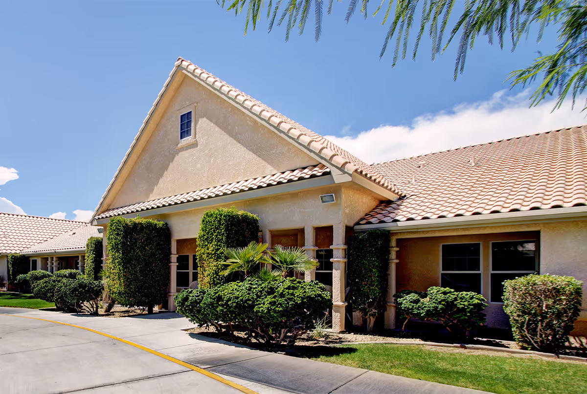 Exterior view of a single-story senior living facility building with beige stucco walls and a tiled roof. The building is surrounded by neatly trimmed bushes and small palm trees under a clear blue sky.
