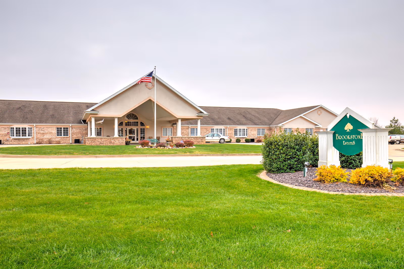 Front of a single-story senior living building with a covered entrance, flagpole, and a green sign on the lawn reading 'Brookstone Estates'.