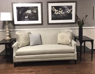 Beige upholstered sofa with patterned pillows between two dark side tables, a lamp and floral artwork on a textured wall.
