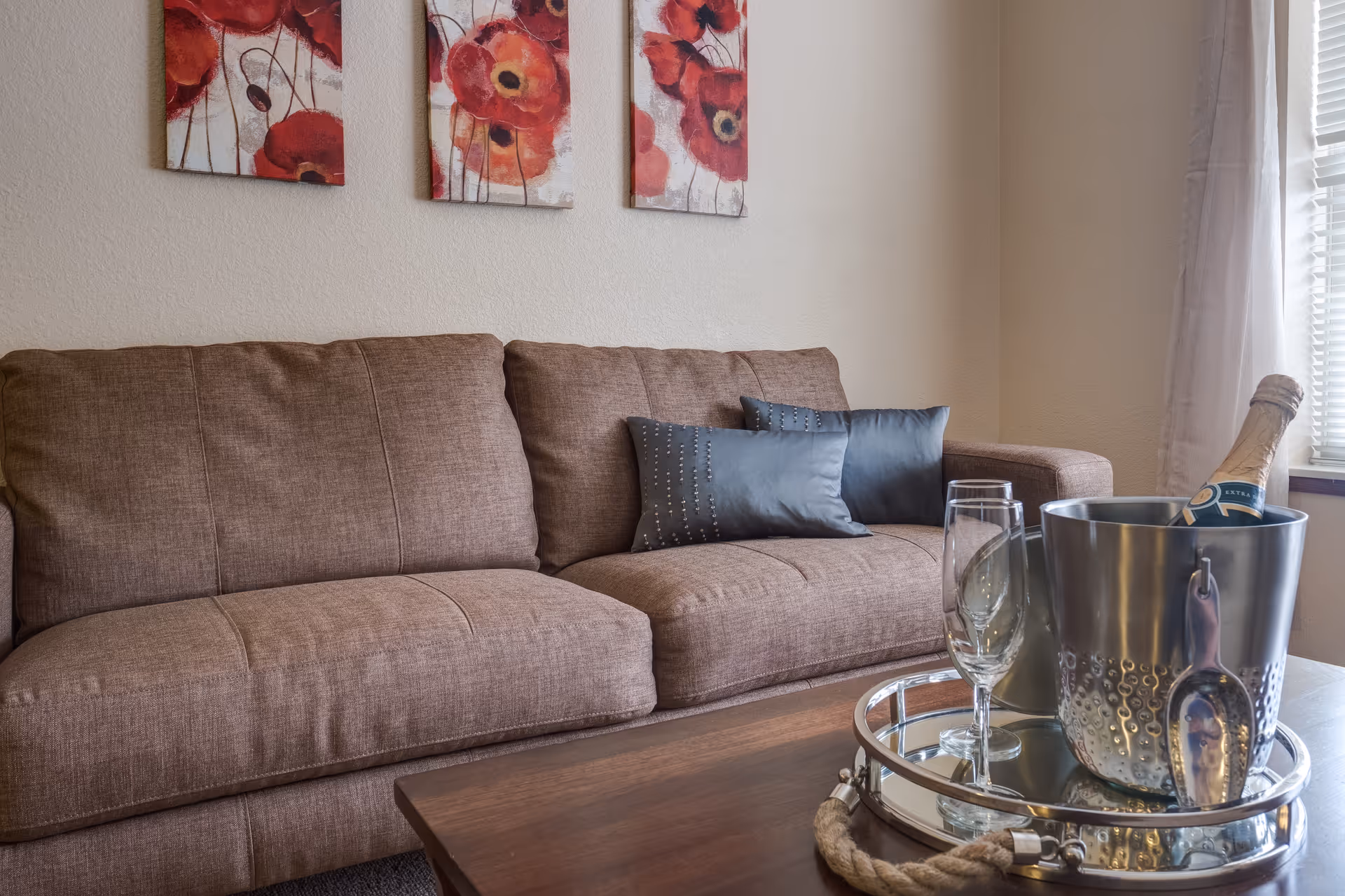 A cozy living room with a brown fabric sofa adorned with two dark gray decorative pillows. Above the sofa, there are three wall art pieces featuring red poppy flowers. In front of the sofa, a wooden coffee table holds a silver tray with a metal ice bucket containing a bottle of champagne and two empty champagne flutes. Light filters in through a window with white blinds and curtains on the right side.