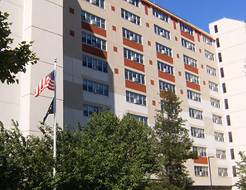 Exterior view of a multi-story senior living facility building with multiple windows, an American flag on a flagpole, and trees in the foreground under a clear blue sky.