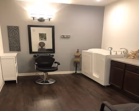 Interior room with a salon chair and mirror next to a walk-in bathtub and sink cabinet on dark wood floors.