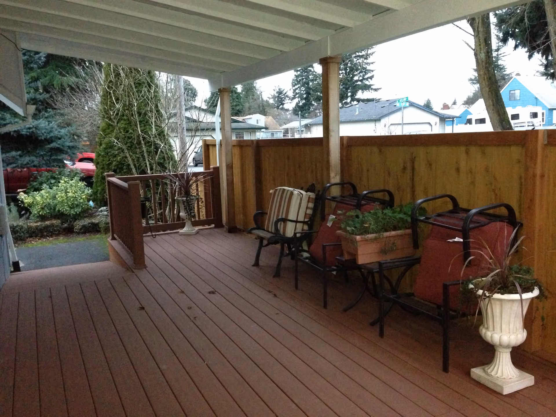 Covered outdoor patio area with wooden flooring and railing, featuring two chairs with cushions, two red carts, and several potted plants. A wooden fence encloses the space, and trees and houses are visible in the background.