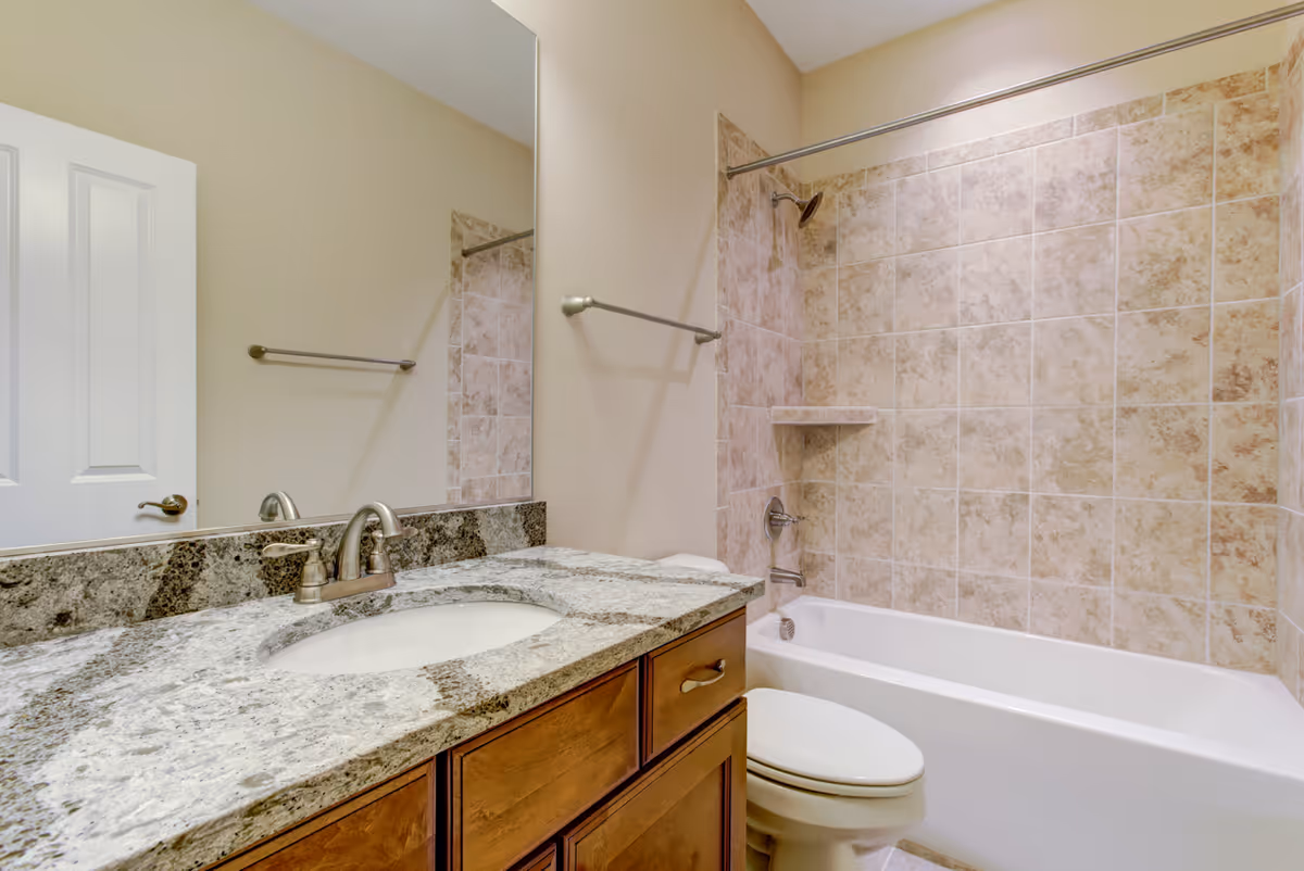 A clean bathroom featuring a granite countertop with an under-mount sink and brushed nickel faucet. There is a large mirror above the sink, a wooden vanity with drawers, a white toilet, and a bathtub with beige tiled walls and a showerhead.