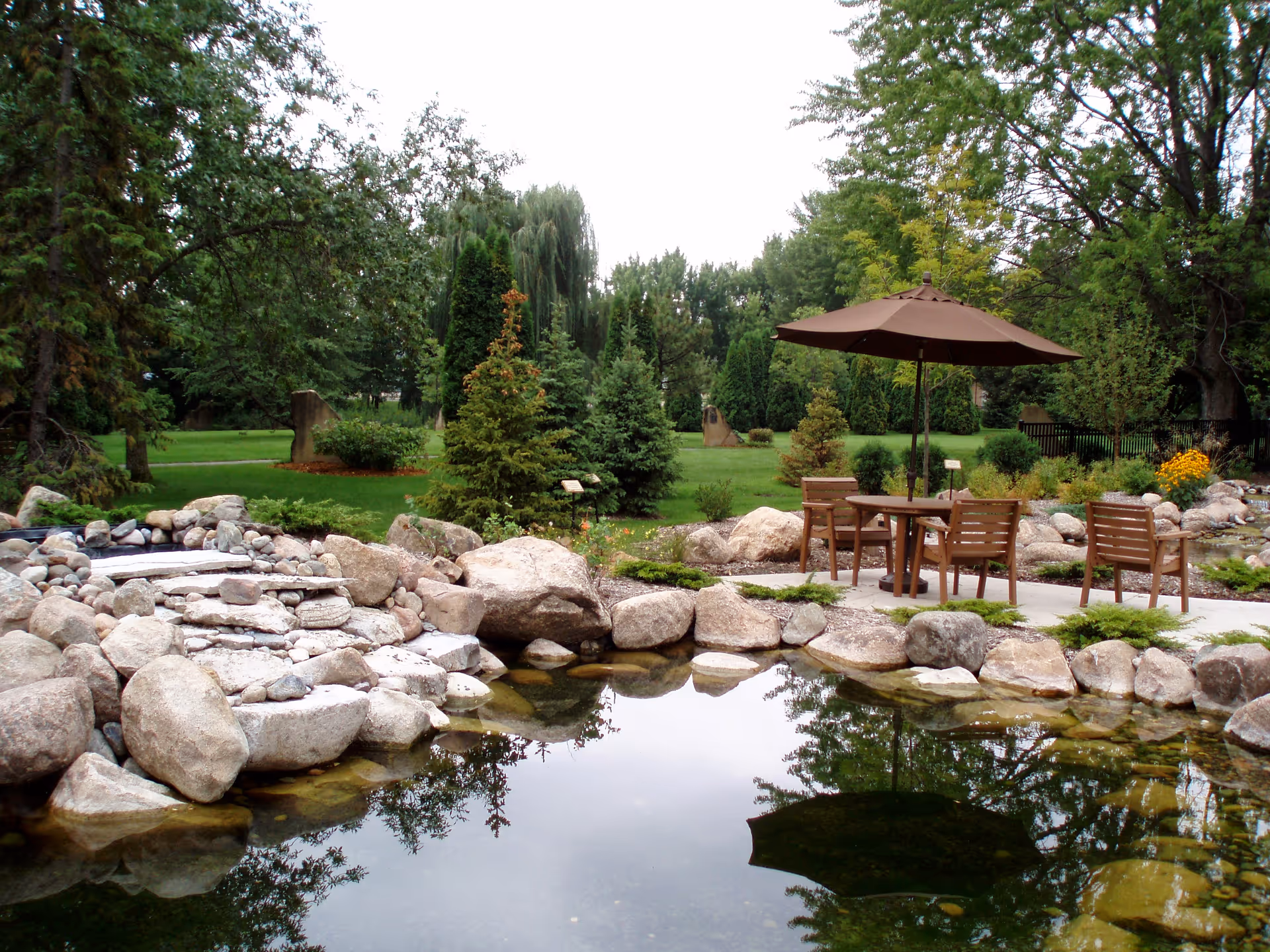 A serene outdoor garden area with a small pond surrounded by large rocks. There is a patio with a round table, four wooden chairs, and a large brown umbrella. The background features lush green grass, various trees, and shrubs.