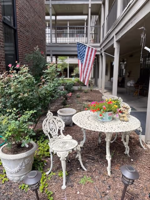 Outdoor patio area with a white ornate metal table and matching chair. On the table is a flower pot with colorful flowers and a small decorative statue. There are potted plants and bushes nearby, with an American flag hanging in the background attached to a building with balconies.