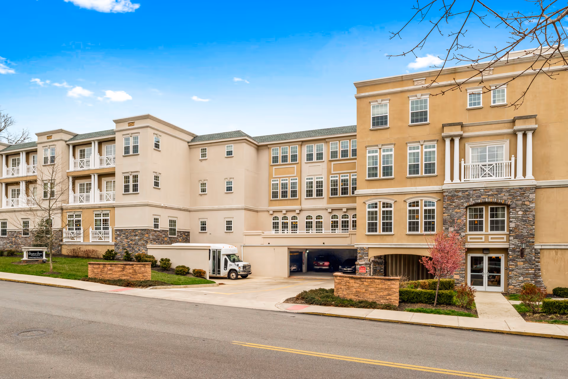 Exterior view of a multi-story senior living facility building with beige and stone facade, multiple windows, balconies, a driveway leading to an underground parking garage, and landscaped areas with small trees and bushes under a clear blue sky.