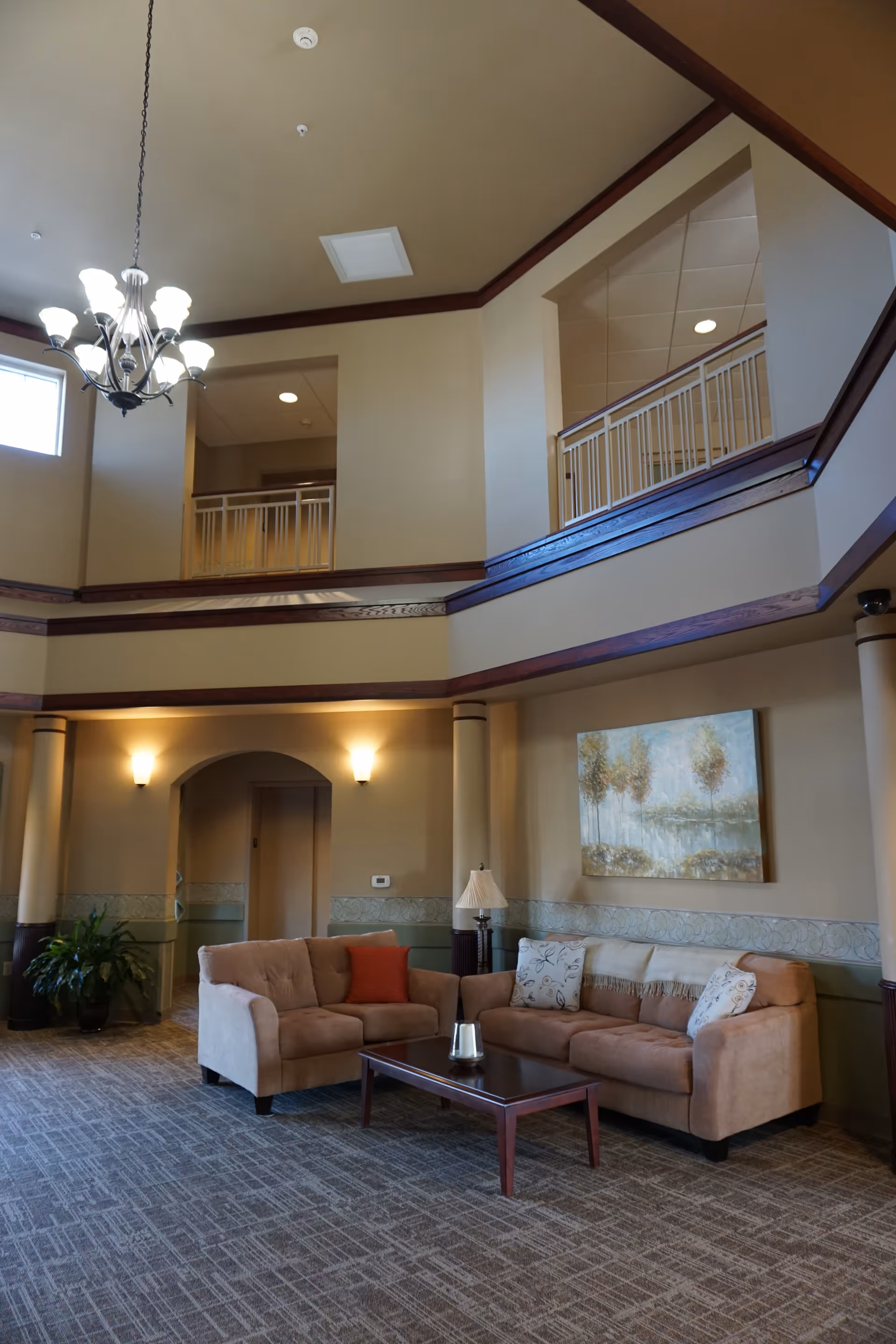 Two-story common lounge with sofas, a coffee table, chandelier, and an upper balcony railing overlooking the seating area.