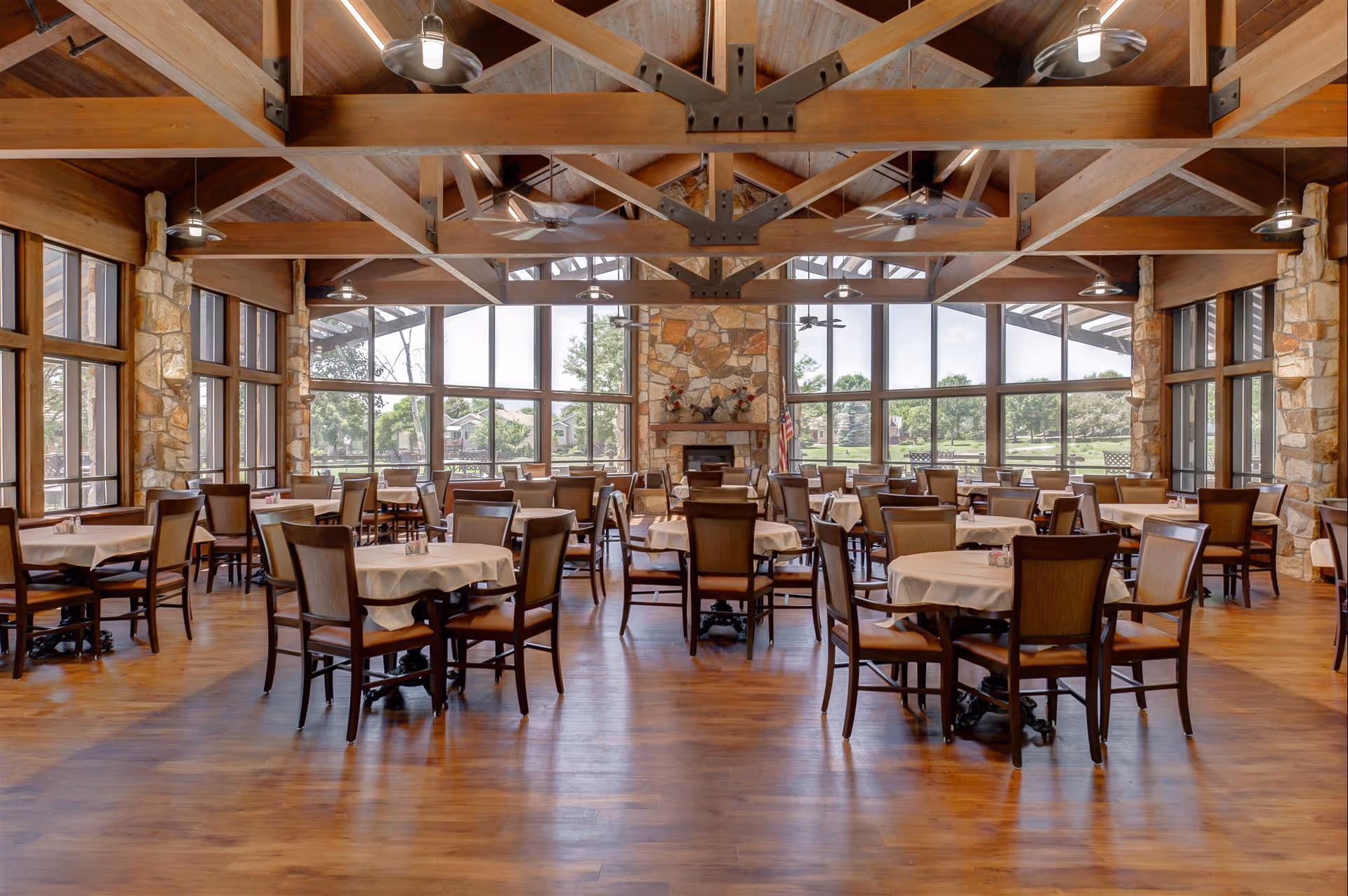 Spacious dining room with multiple round tables covered with white tablecloths and surrounded by wooden chairs. The room features large windows allowing natural light to fill the space, wooden beams on the ceiling, ceiling fans, and a stone fireplace at the far end.