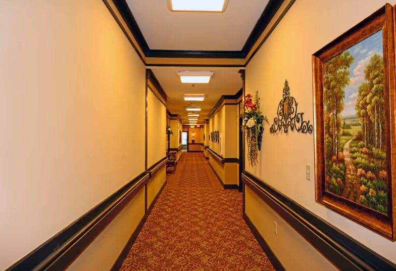 A long, well-lit hallway in a retirement center with beige walls, dark trim, and a patterned carpet. The hallway is decorated with framed paintings and wall-mounted floral arrangements.