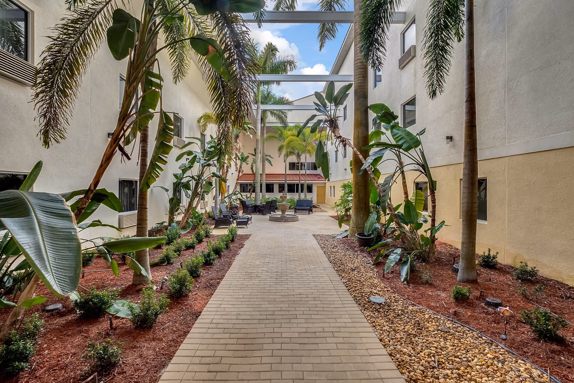 Outdoor courtyard area in an assisted living facility with a paved walkway lined by tropical plants and palm trees. There are seating areas with chairs and a fire pit in the background, surrounded by the building's beige and white walls under a partly cloudy sky.