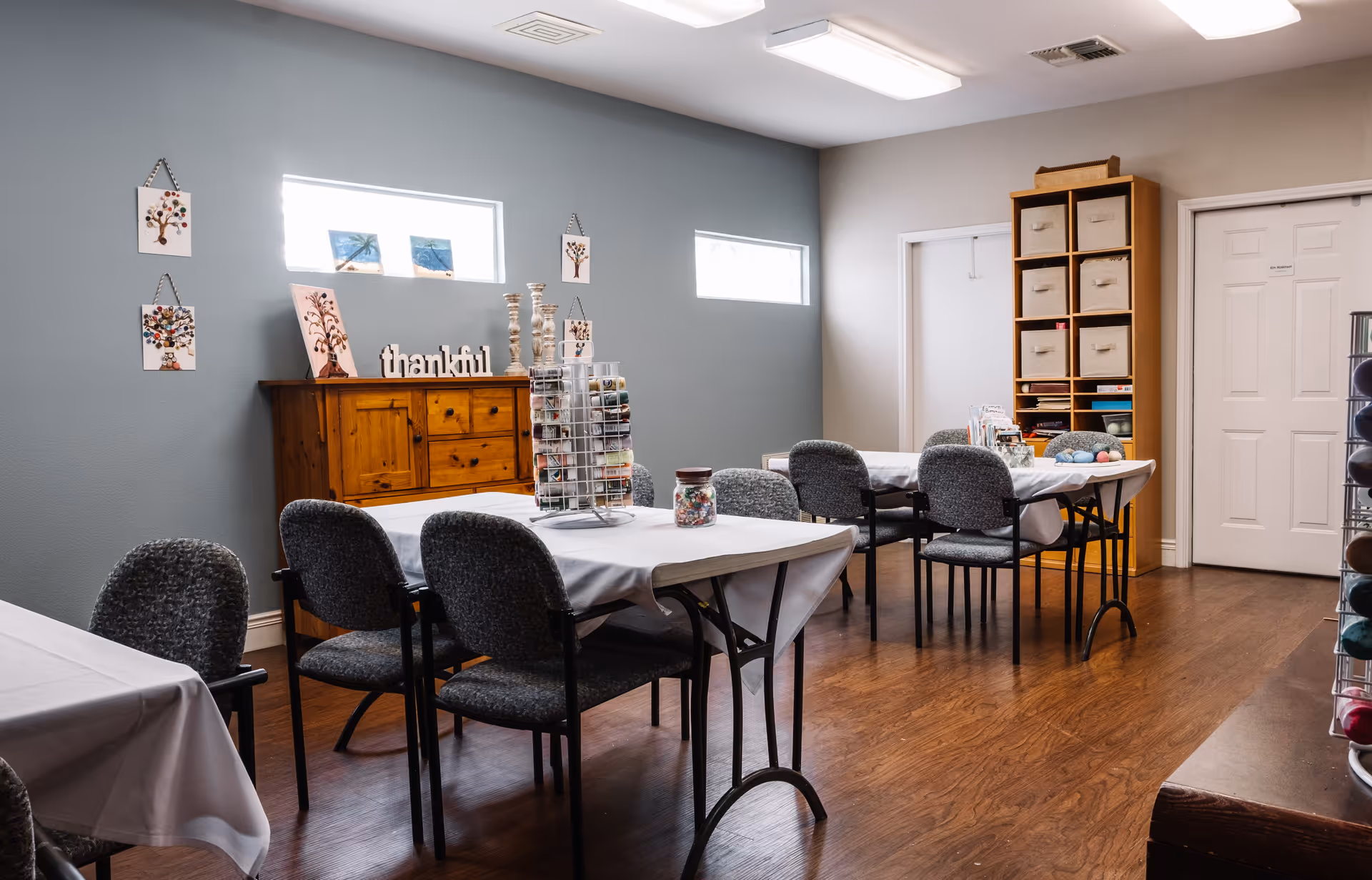 A well-lit room with several tables covered in white tablecloths and surrounded by gray cushioned chairs. The room has wooden flooring, a wooden cabinet against a blue-gray wall with decorative items including a 'thankful' sign, and small artworks hanging on the wall. There are two small rectangular windows letting in natural light. Shelves with storage bins and craft supplies are visible, suggesting the room is used for activities or crafts.
