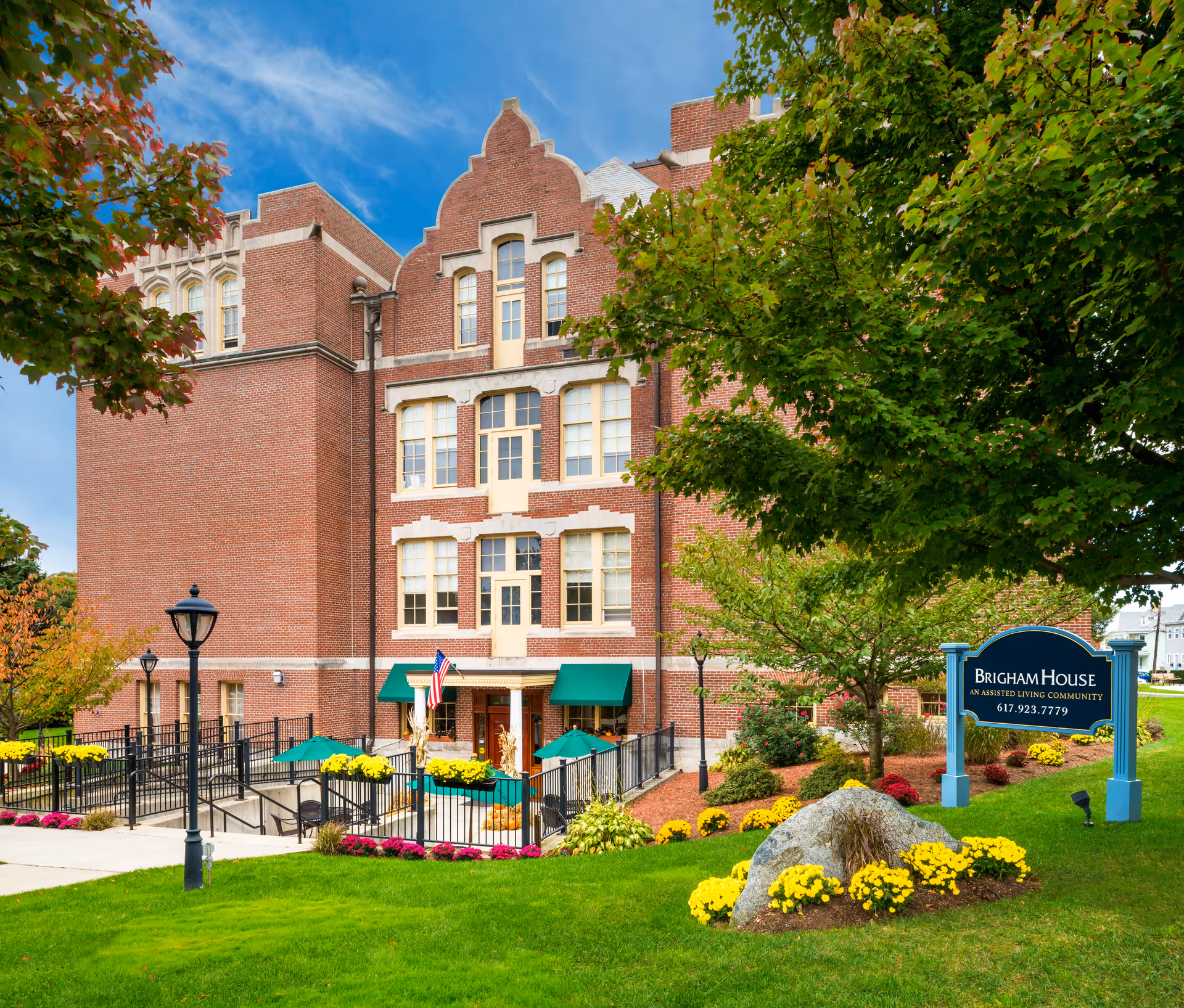 Exterior view of Brigham House, a multi-story brick building with large windows and green awnings. The building is surrounded by well-maintained landscaping including green grass, colorful flowers, and trees. There is a blue sign in the foreground that reads 'Brigham House, An Assisted Living Community, 617.923.7779'. The sky is clear and blue.