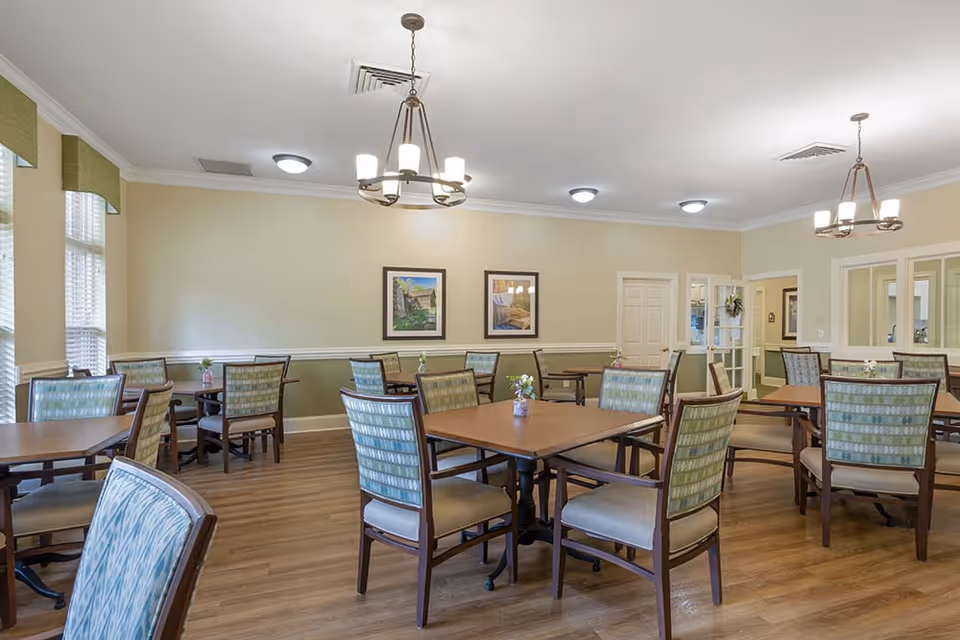 A bright dining room with multiple wooden tables and cushioned chairs arranged neatly. The room has light-colored walls with two framed pictures, large windows with green valances, and two chandeliers hanging from the ceiling. Small flower vases are placed on each table.
