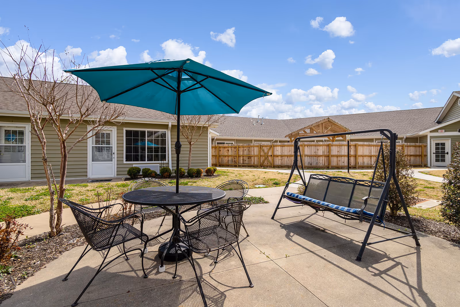 Outdoor patio area at Brookfield Assisted Living and Memory Care featuring a round metal table with four matching chairs and a large teal umbrella. Nearby is a metal swing bench with a blue and white cushion. The patio is surrounded by a concrete walkway, small bushes, and a wooden fence. The building exterior is visible with beige siding, white doors, and windows under a partly cloudy blue sky.