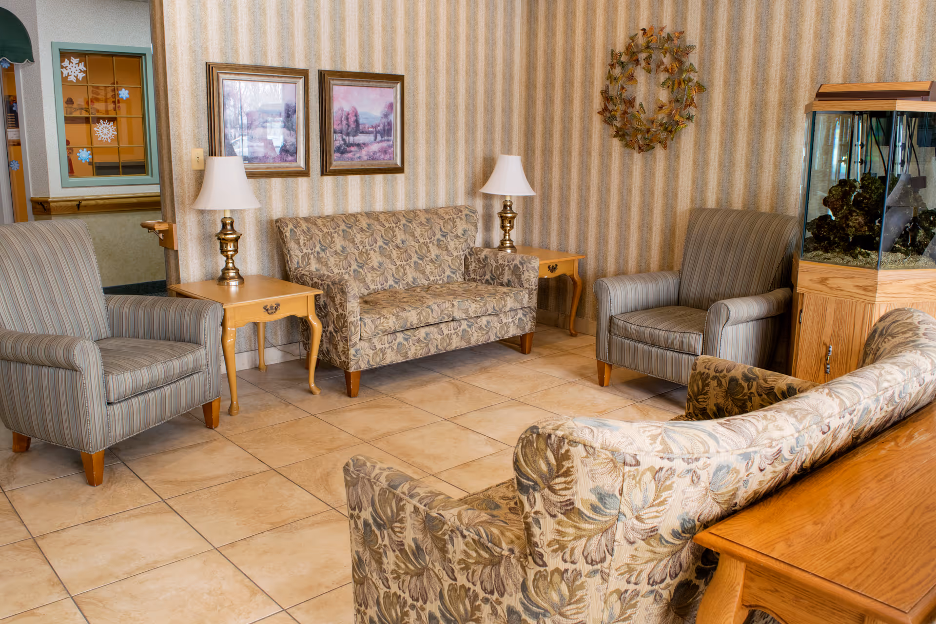 A cozy assisted-living lounge with patterned sofas and armchairs arranged around side tables and lamps, with an aquarium on the right.