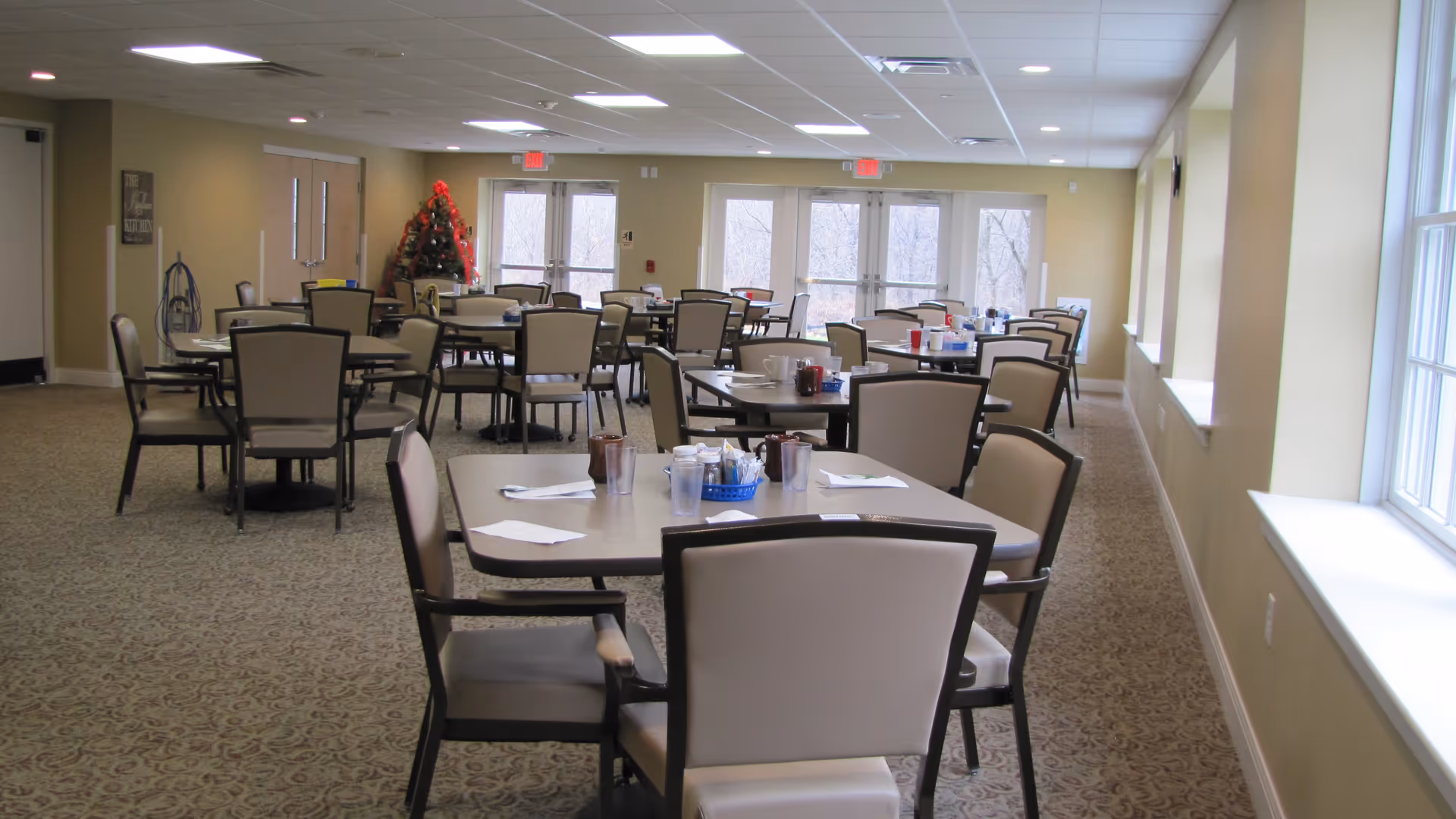 A spacious dining room with multiple tables and chairs arranged neatly. Each table has condiments and napkins placed on it. Large windows on the right side allow natural light to fill the room. There is a Christmas tree decorated in the back corner near double doors.