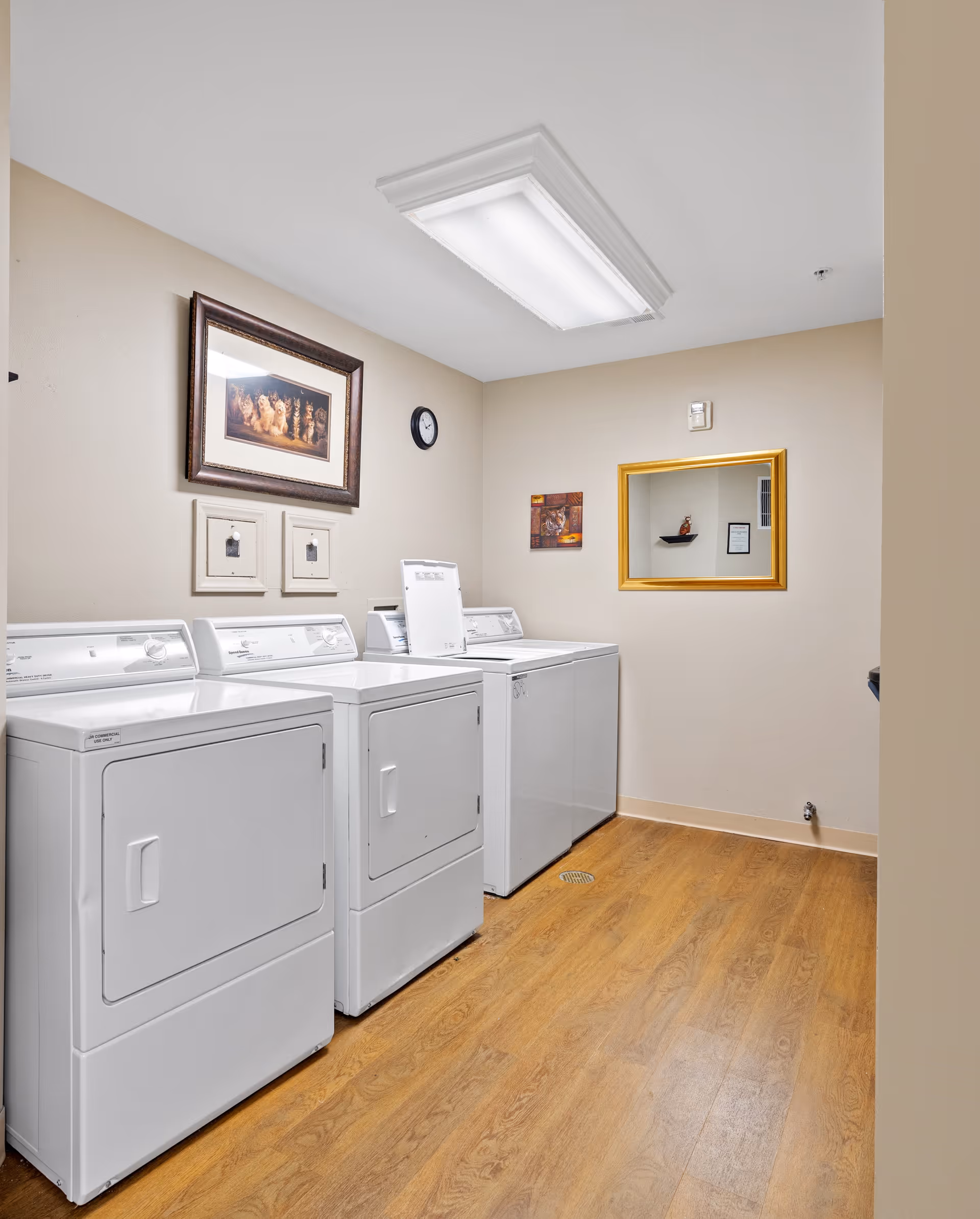 Laundry room with three white washing machines and dryers lined up against a beige wall. The room has wood flooring, a large rectangular ceiling light, a wall clock, a framed picture, two small framed artworks, and a gold-framed mirror on the walls.