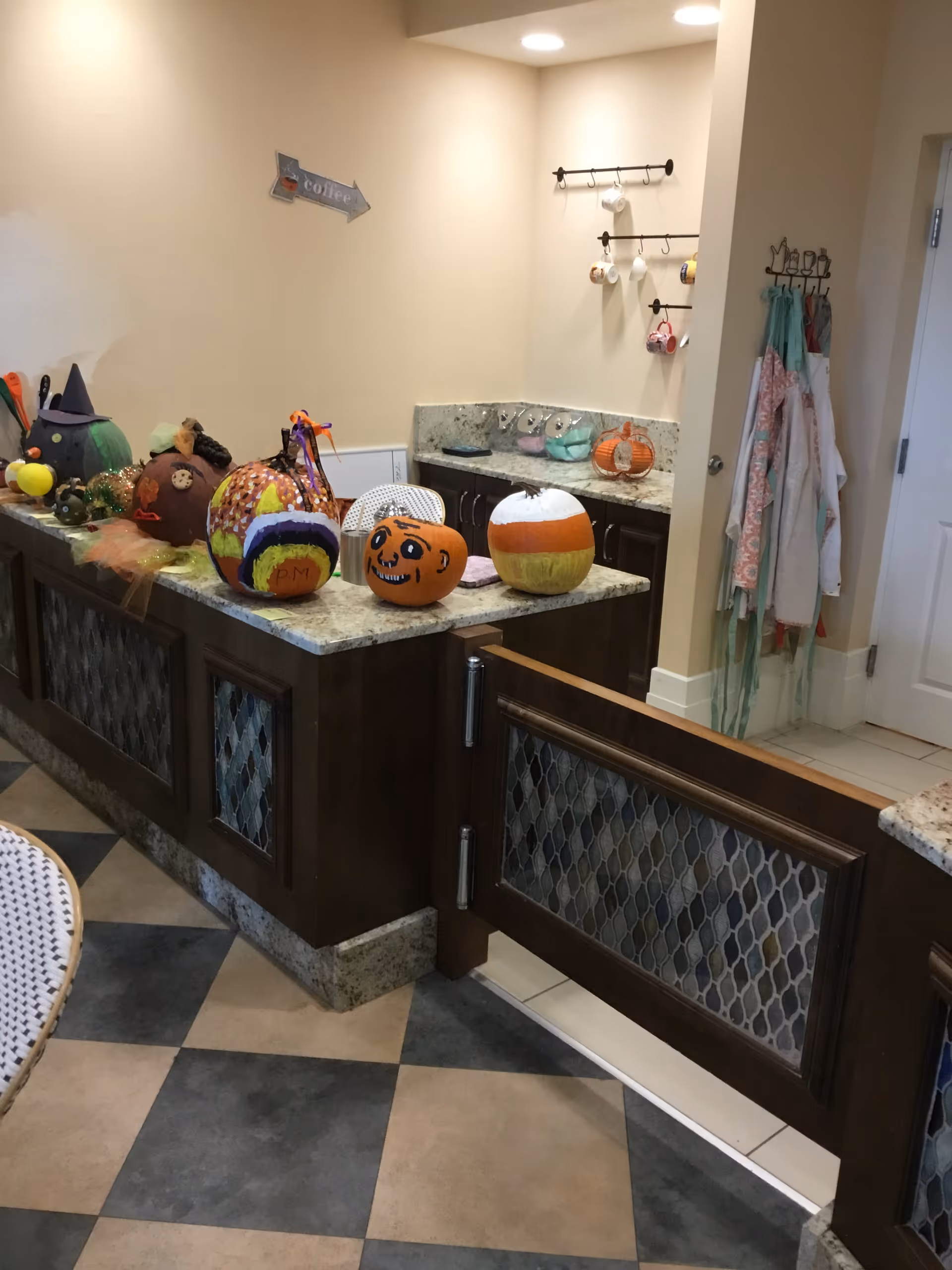 Interior view of a kitchen area with a granite countertop island decorated with various painted and decorated pumpkins. The kitchen has dark wood cabinets, a tiled floor with a checkered pattern, and hooks on the wall holding mugs and aprons. A small wooden gate is attached to the island.