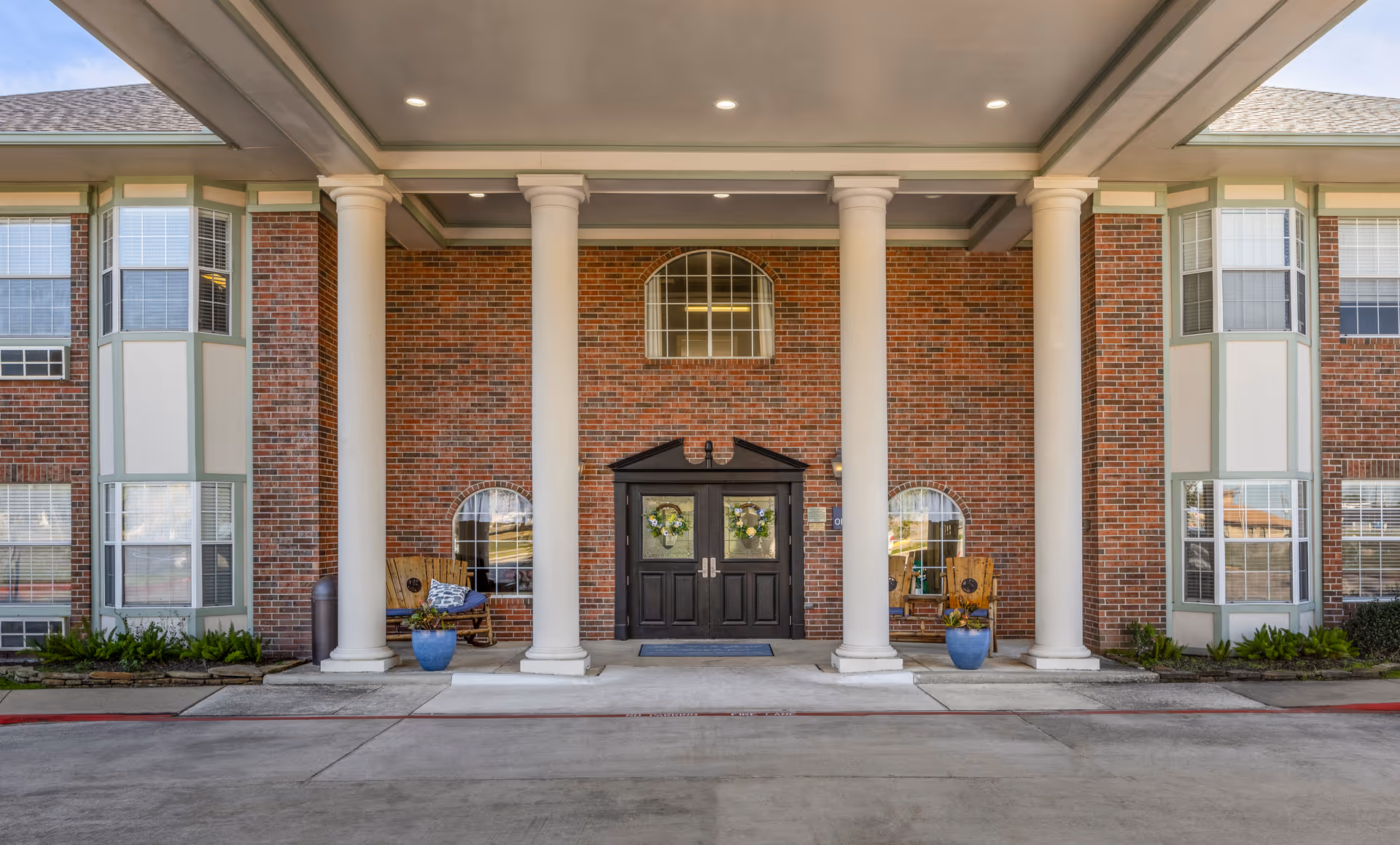 Front entrance of a brick building with four large white columns supporting a covered porch. The entrance has double black doors decorated with wreaths, flanked by two wooden chairs with cushions and blue flower pots. The building has multiple windows and a concrete driveway in front.