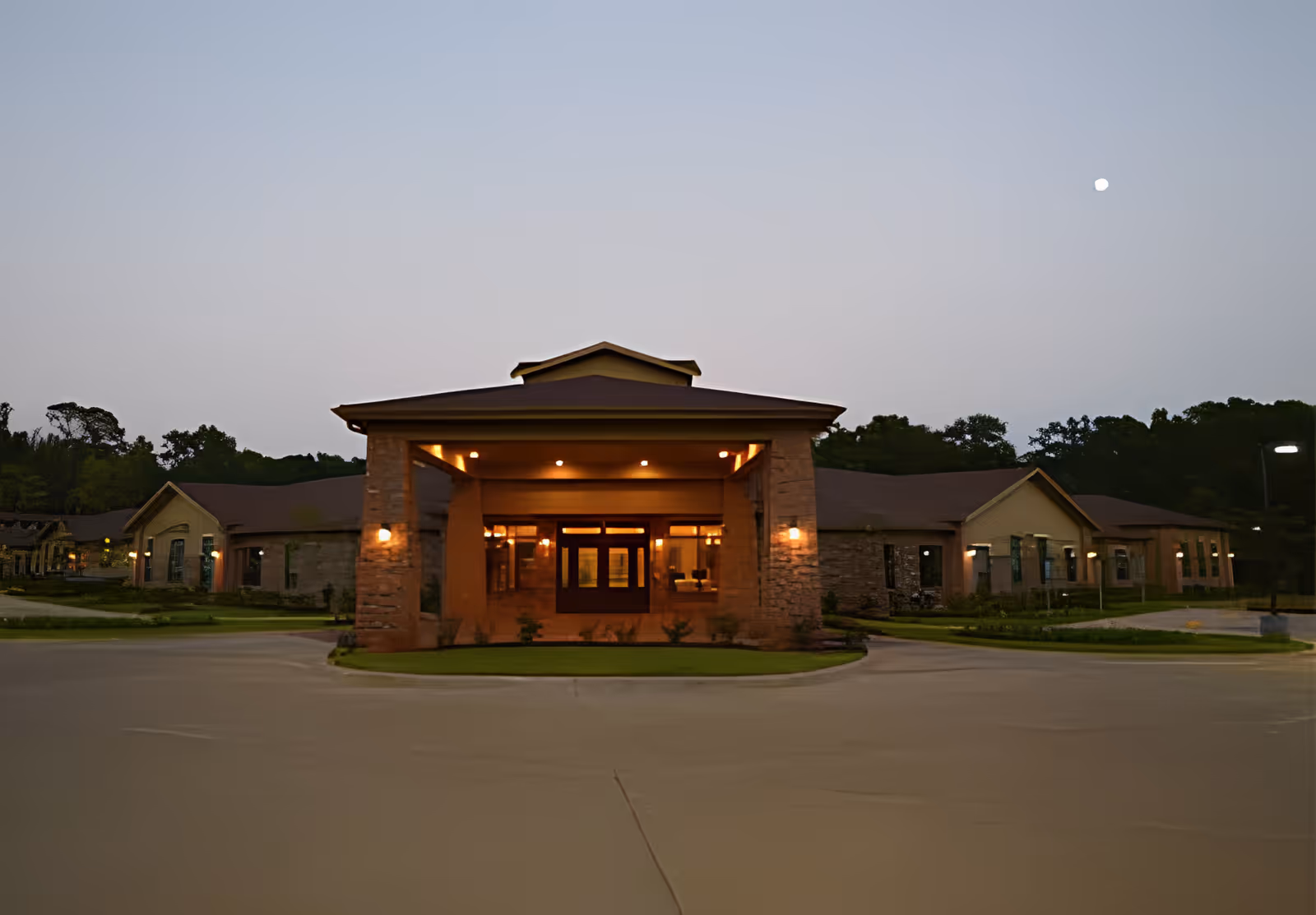 Exterior view of Windermere at Cartmell facility at dusk, showing the main entrance with stone pillars and warm lighting, surrounded by a driveway and landscaped greenery.