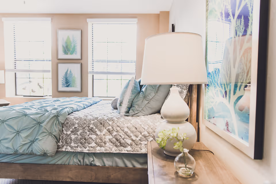 Bright bedroom with a made bed, blue-green bedding, a bedside lamp and framed artwork.
