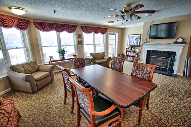 A cozy living room area with a rectangular wooden table surrounded by six chairs with floral upholstery. There are two armchairs near large windows with red valance curtains. A fireplace with a mounted flat-screen TV above it is on the right wall, next to a bookshelf and a framed picture. The room has carpeted flooring and ceiling fans with lights.
