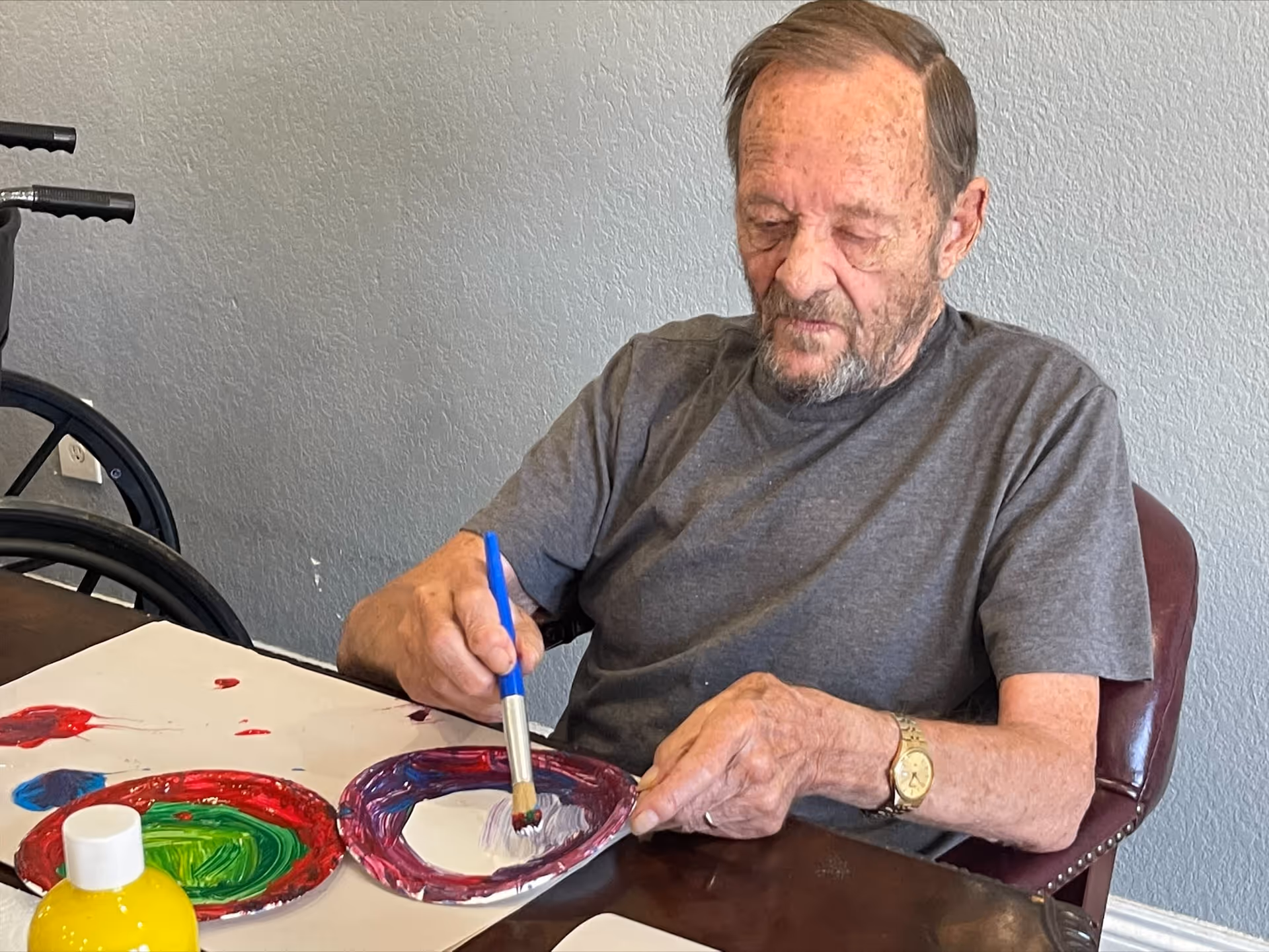 An elderly man sitting at a table painting on paper with a paintbrush. There are colorful paint plates and a bottle of yellow paint on the table. A wheelchair is visible in the background against a gray wall.