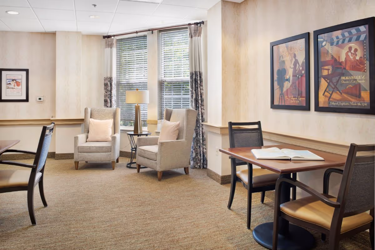 Sunlit communal lounge with two upholstered armchairs by a window, a side table and lamp, and a small table with chairs and framed artwork on the wall.