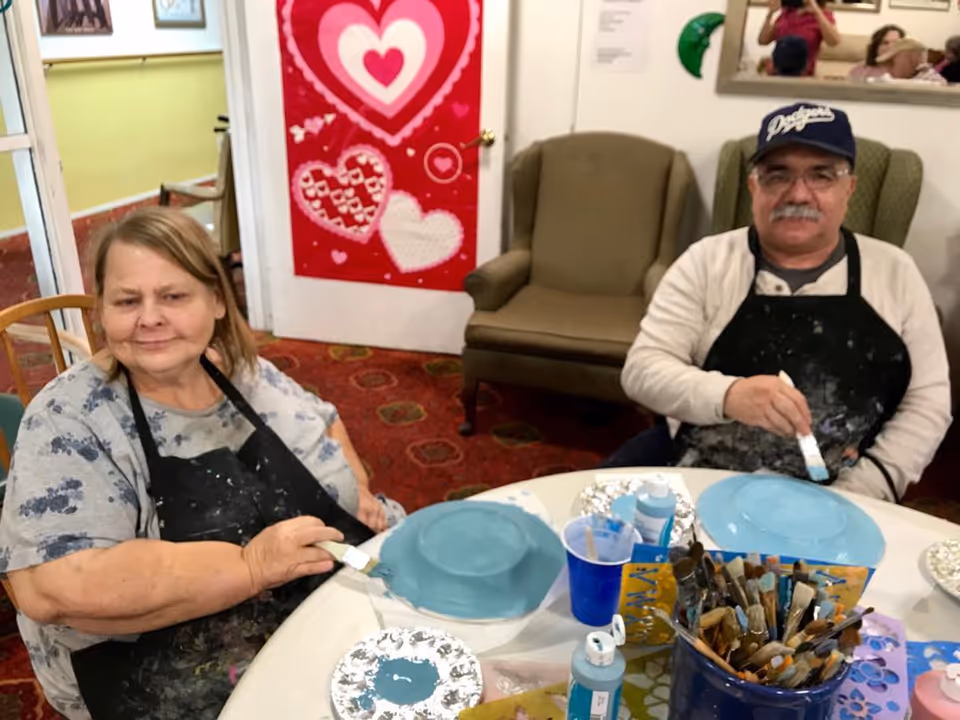 Two elderly individuals sitting at a round table engaged in painting crafts. Both are wearing aprons and holding paintbrushes, painting blue on round objects. Behind them is a wall with a red decoration featuring hearts and a green armchair.