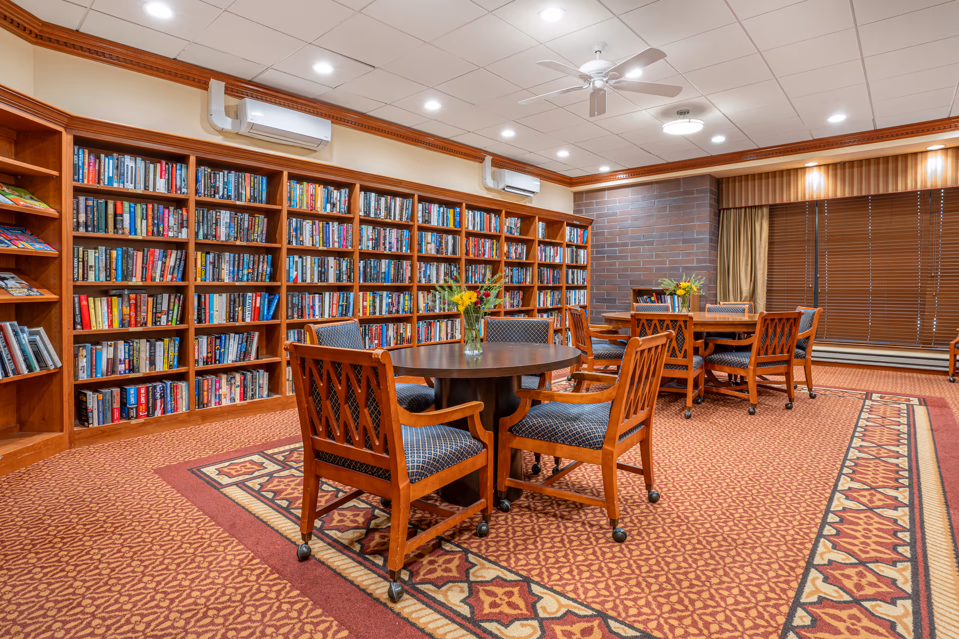 A cozy library room with wooden bookshelves filled with books along the wall. There are round wooden tables with cushioned chairs on wheels arranged on a patterned carpet. The room has ceiling lights, a ceiling fan, and large windows with closed blinds and curtains.
