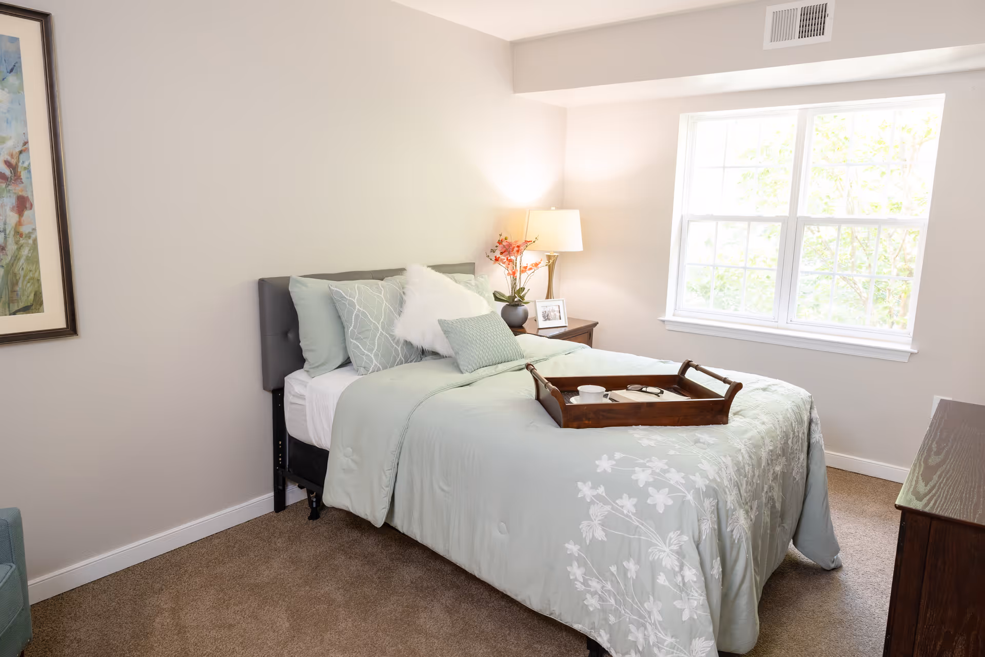 Bright bedroom with a neatly made bed in pale green bedding, a wooden tray on the bed, a bedside lamp and a large window.
