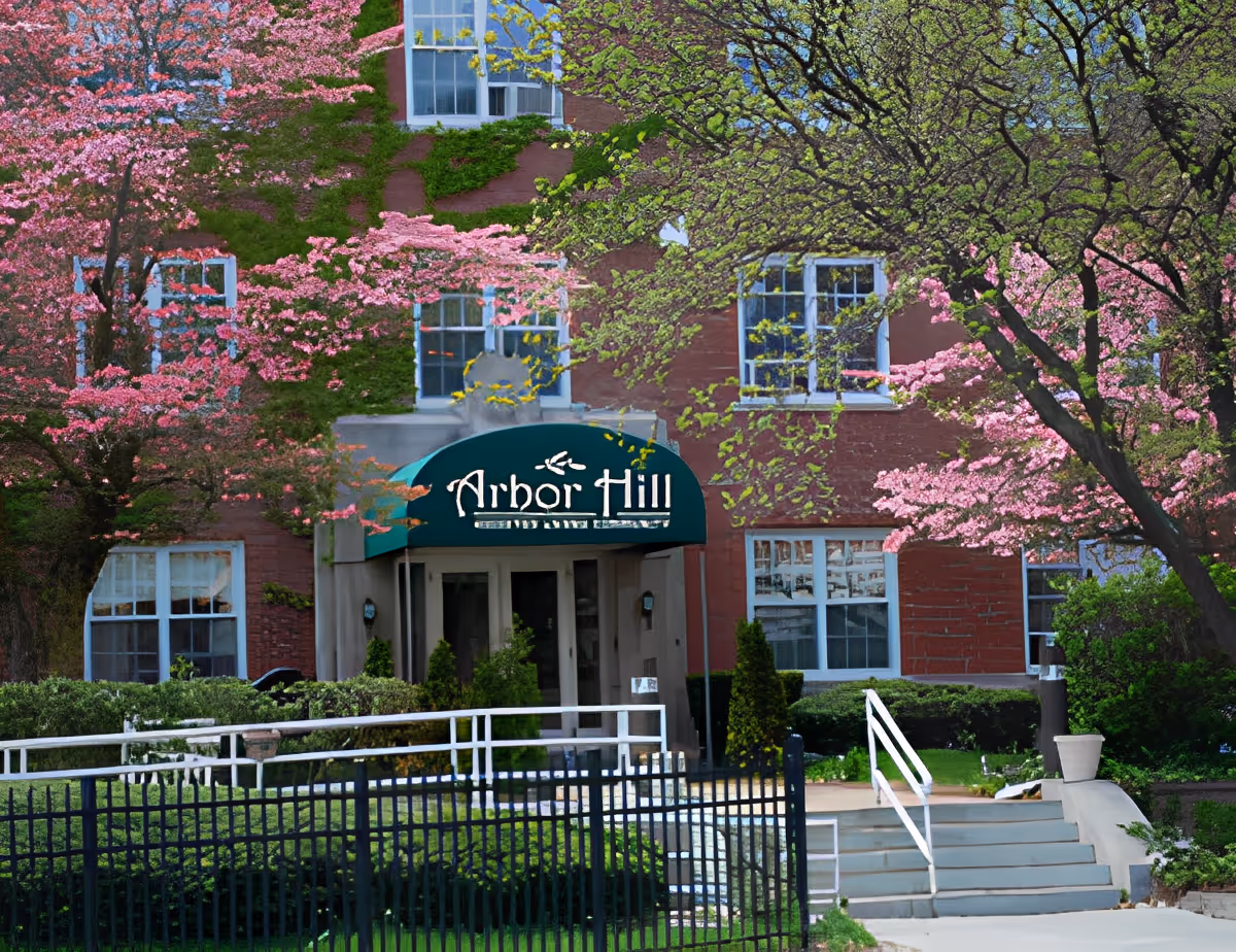 Exterior view of Arbor Hill Assisted Living facility with a brick building partially covered in green ivy. The entrance has a green awning with the facility name. Surrounding the building are trees with pink blossoms and green leaves, along with bushes and a black metal fence in front.