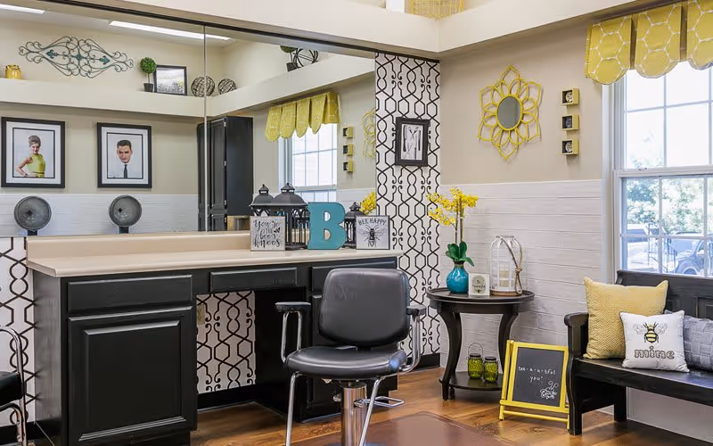 A cozy interior space with a black salon chair in front of a counter with decorative items including lanterns, a blue letter B, and small signs. The walls have a patterned wallpaper and a large mirror reflecting the room. There is a small round table with a blue vase holding yellow flowers, a lantern, and a small framed picture. A black bench with yellow and white pillows sits near a window with yellow valances. The room has a warm and inviting atmosphere with wood flooring.