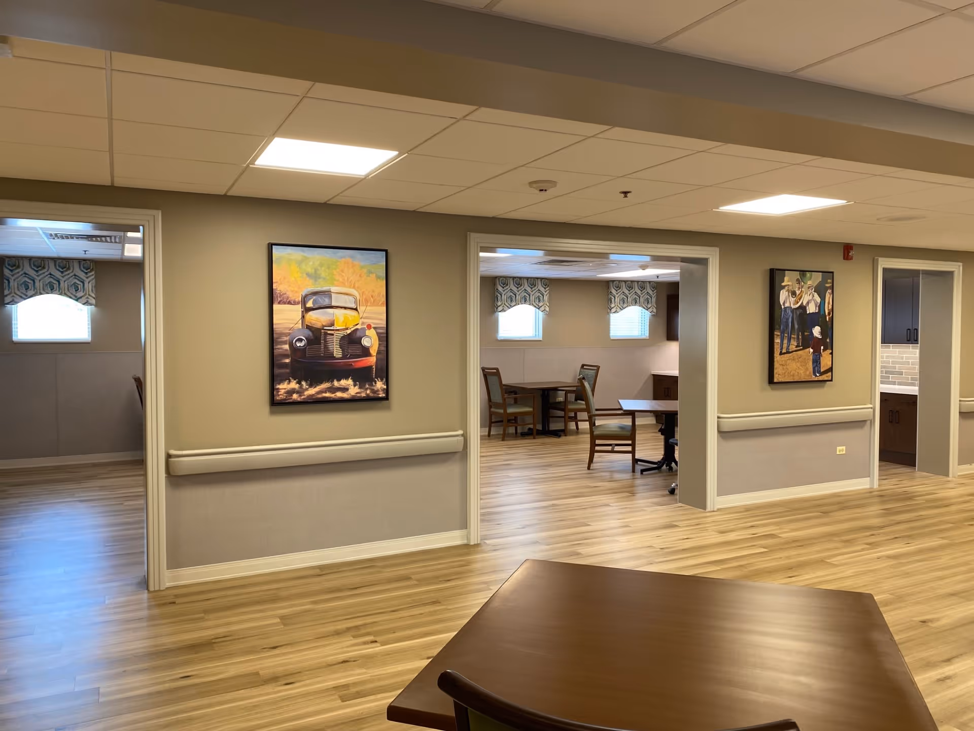 Interior view of a senior living facility showing a hallway with light wood flooring and beige walls. There are three open doorways leading to rooms with tables and chairs. Two framed paintings hang on the walls, one depicting a vintage car and the other showing people in a rural setting. The ceiling has recessed lighting panels, and windows with patterned valances provide natural light in the rooms.
