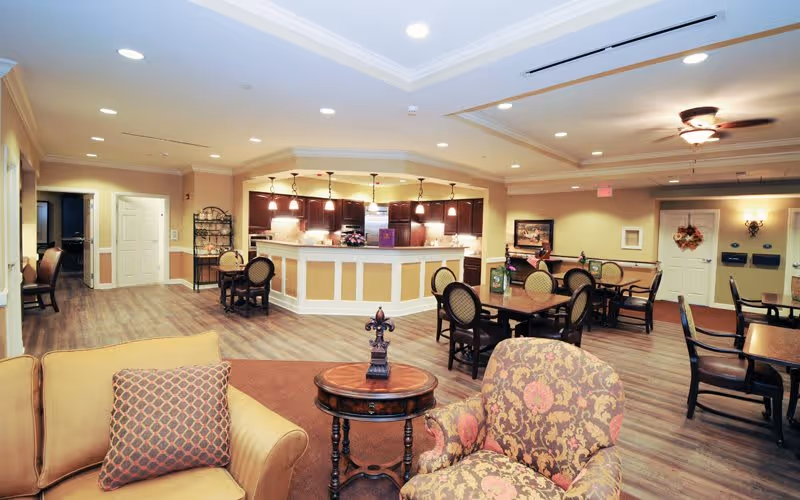 A spacious and well-lit common area in a senior living facility featuring a seating area with upholstered chairs and a small round wooden table in the foreground. In the background, there is a large counter with pendant lighting above, several dining tables with chairs, and warm beige walls with decorative accents. The floor is wood-style laminate, and the ceiling has recessed lighting and a ceiling fan.