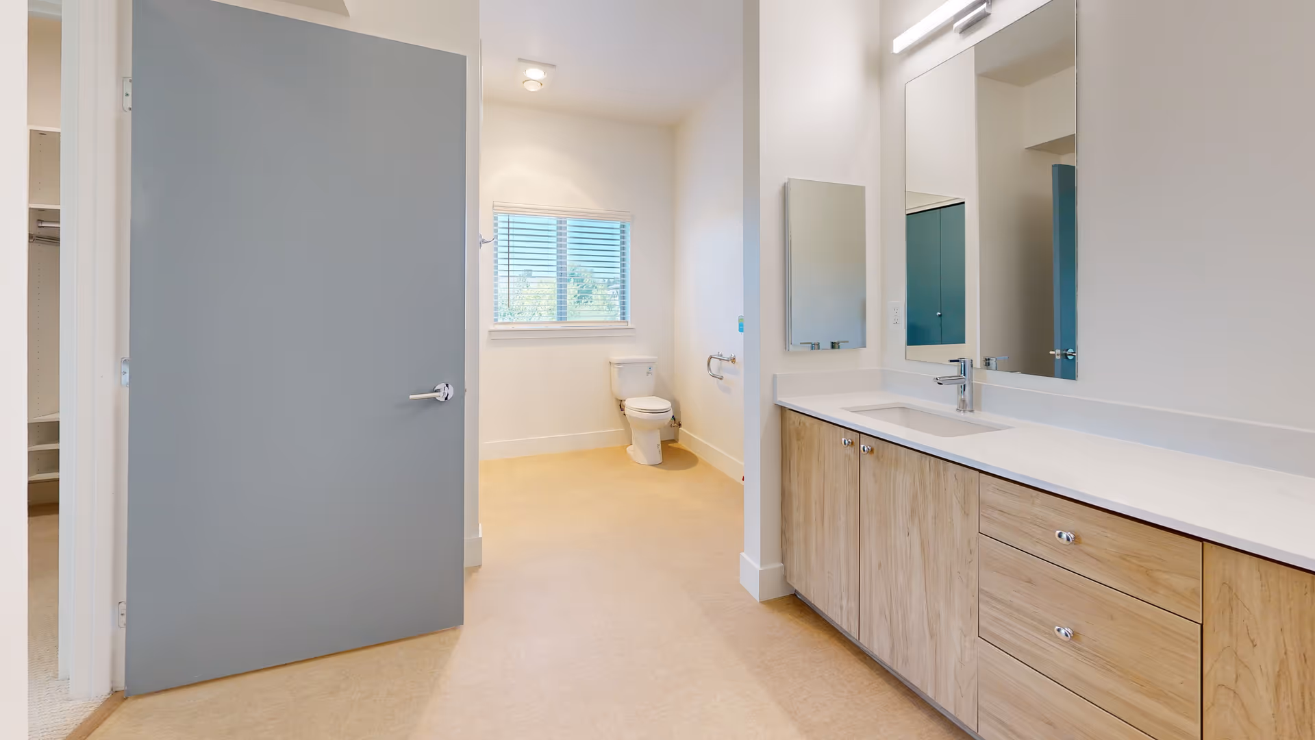 Bright bathroom with a large vanity featuring a white countertop and wooden cabinets, a rectangular mirror above the sink, a toilet near a window with blinds, and a partially open gray door leading to another room.
