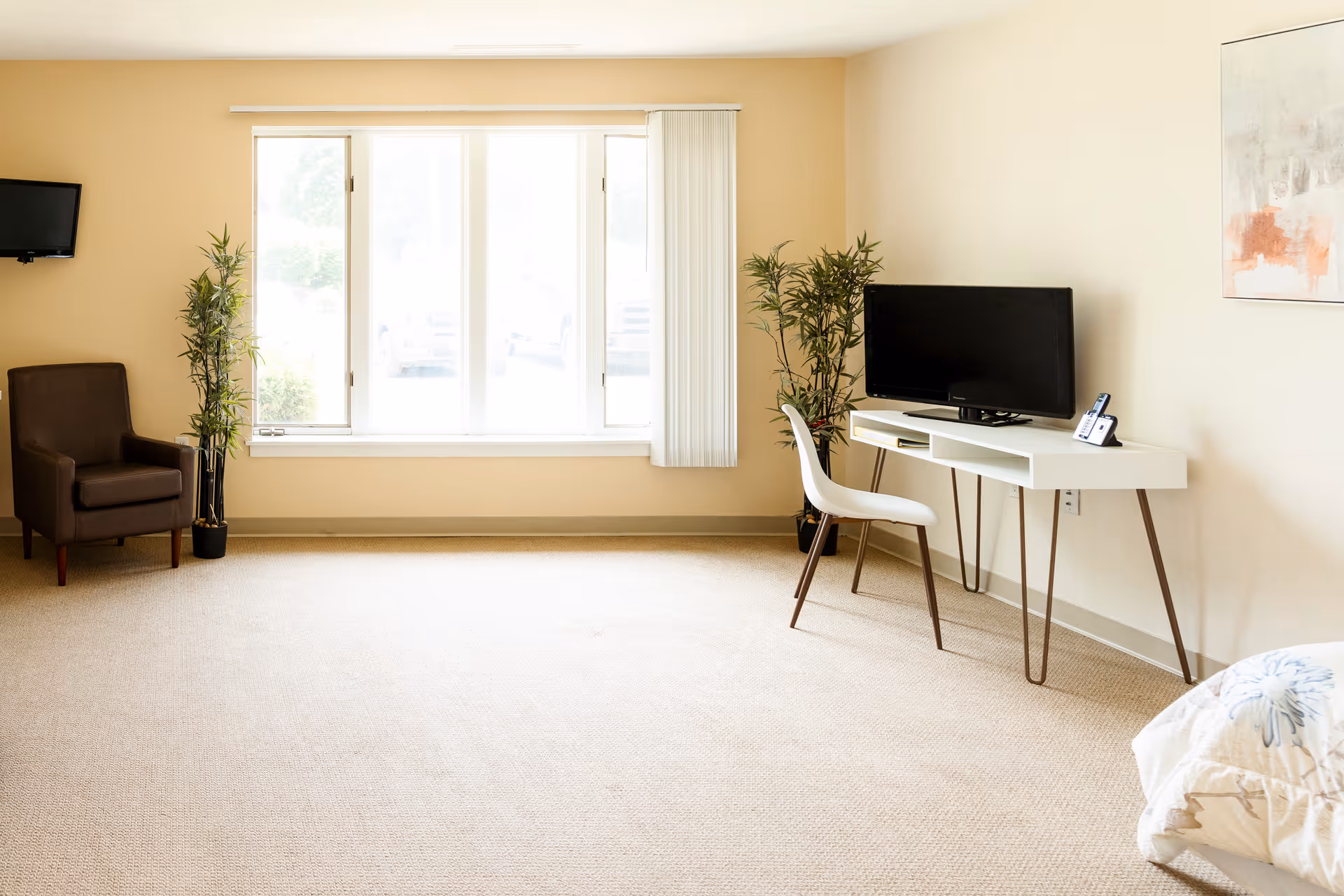 Bright, sparsely furnished living room with a large window, armchair, desk with a television and a chair.