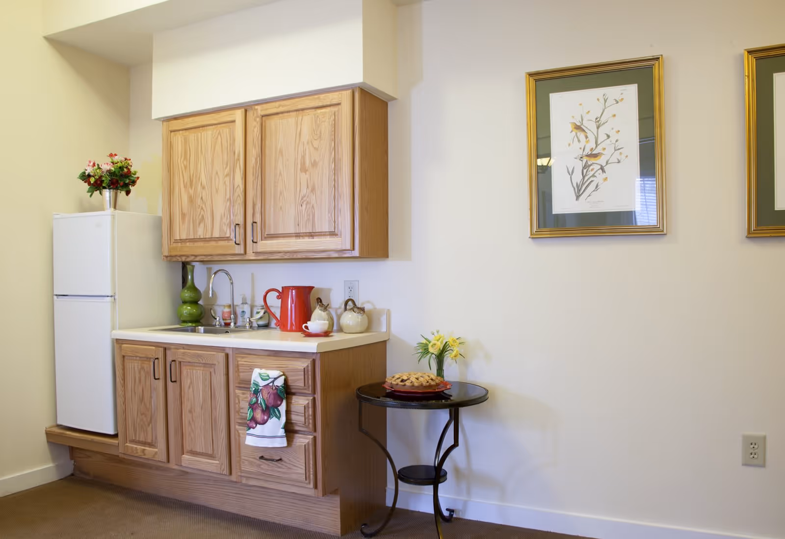 Small kitchenette area with wooden cabinets, a white refrigerator, a sink, and a countertop holding a red pitcher, cups, and decorative items. A small round table next to the kitchenette has a pie and a vase with yellow flowers. Two framed bird illustrations hang on the wall above the table.