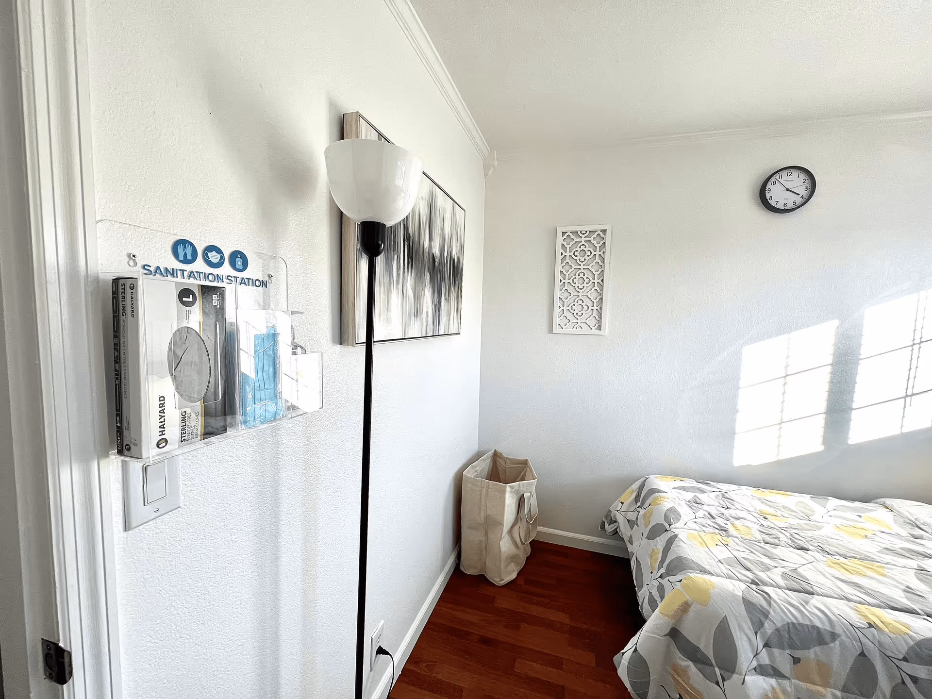 Sunlit small bedroom with a bed, floor lamp, wall art and a mounted sanitation station by the door.